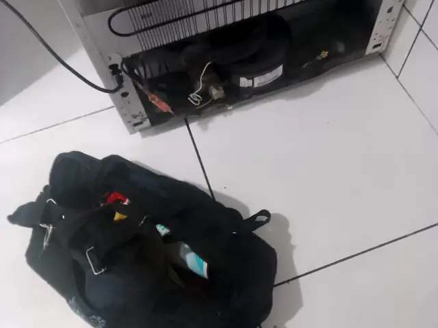 Geladeira vista por trás, com o compressor à mostra e a bolsa de ferramentas no chão
