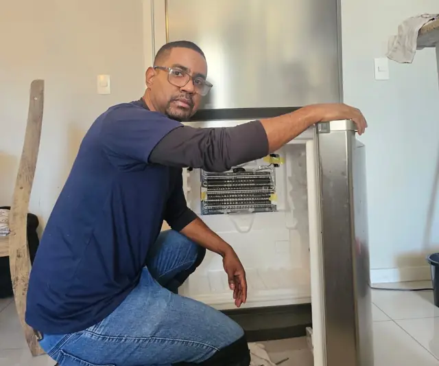Luiz, técnico da Luitec, agachado ao lado de uma geladeira Brastemp aberta, durante o conserto